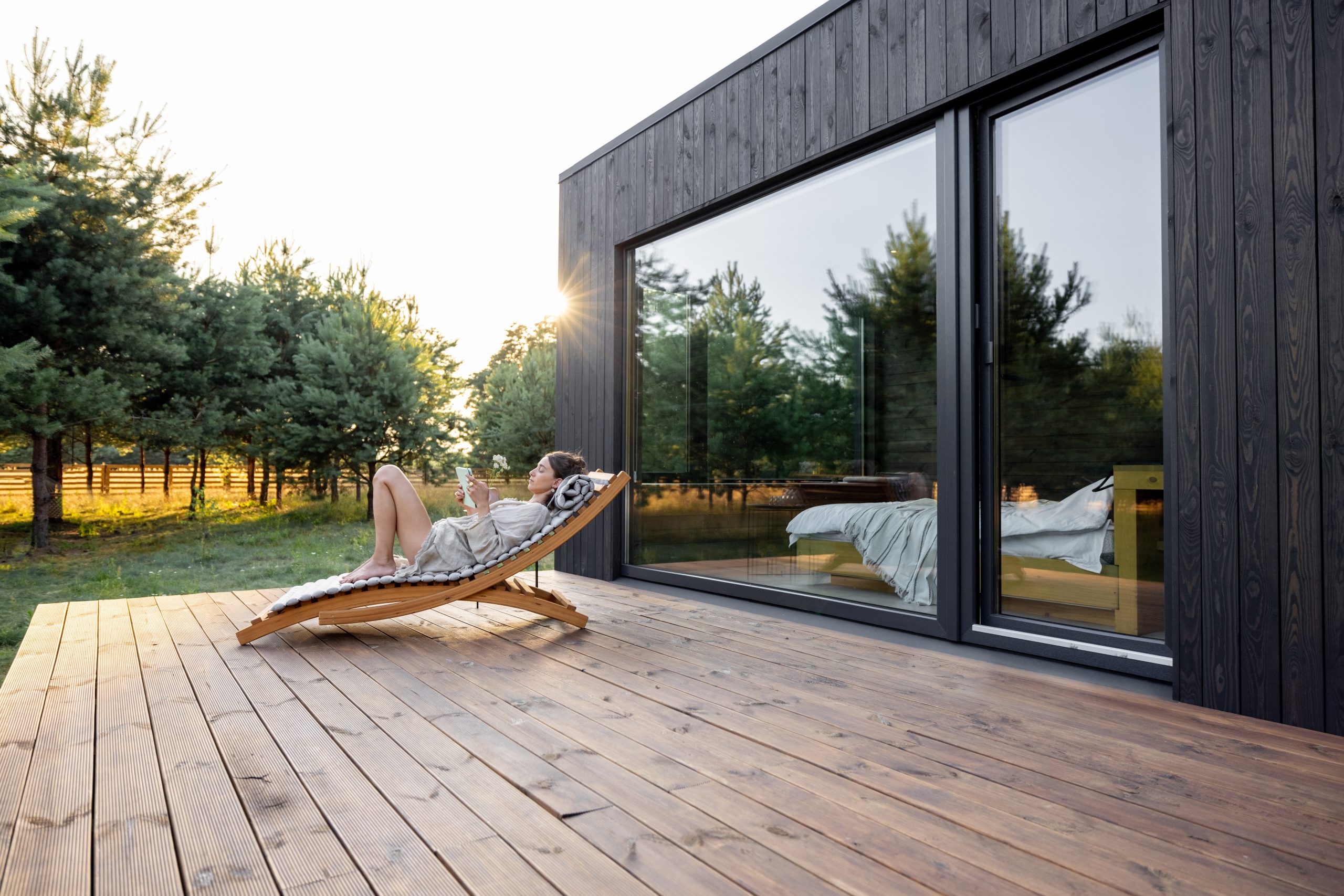 Eine Frau entspannt sich in einem modernen Garten auf einer Holzterrasse eines zeitgemäßen schwarzen Hauses, während sie auf einem stilvollen Liegestuhl in der Sonne liest. Der Hintergrund zeigt eine ruhige Umgebung mit Bäumen und einem grünen Feld, was die harmonische Integration von Architektur und Natur betont.